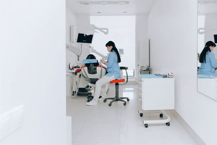 A dentist in a blue coat and mask works in a bright, modern dental office.