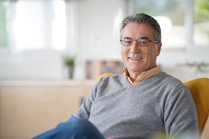 Older man with glasses and grey shirt sitting in chair and smiling.